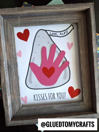 Framed craft featuring a Hersheys Kiss drawing, decorated with red and pink hearts and a pink paper handprint—perfect as a Mother's Day keepsake. A banner says Love, Alex. Text at the bottom reads Kisses for You! and @GLUEDTOMYCRAFTS is at the bottom.