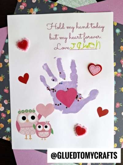 A handmade Valentine’s card doubles as a Mother’s Day keepsake, featuring a purple handprint, red and pink felt hearts, cartoon owls, and floral background. The text reads, “Hold my hand today but my heart forever. Love Jason.”.