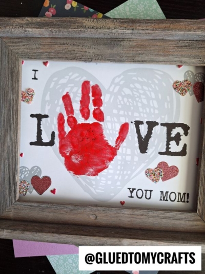 A framed mother's day keepsake with I LOVE YOU MOM! on white paper. The O in LOVE is replaced by a red child's handprint, surrounded by decorative hearts. The bottom features the handle @GLUEDTOMYCRAFTS.