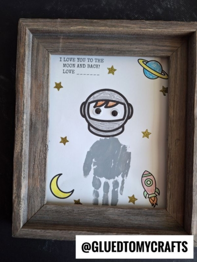 Framed children’s craft with a gray handprint turned into an astronaut, surrounded by stars, a planet, a moon, and a rocket. A perfect Mother’s Day keepsake with text: I love you to the moon and back! Love ______.