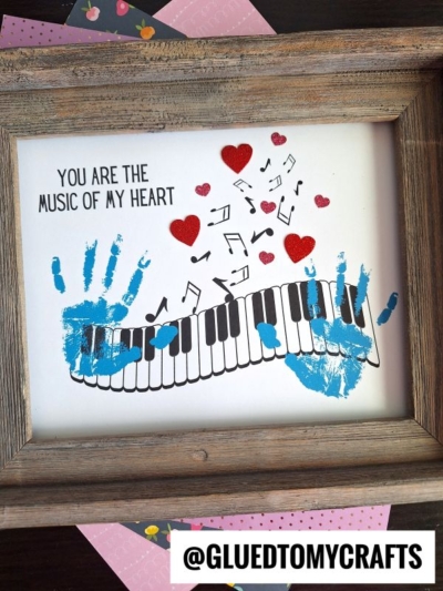 Framed artwork with blue handprints forming a piano keyboard. Red hearts and black music notes float above. A perfect mother’s day keepsake featuring the text, “YOU ARE THE MUSIC OF MY HEART.” @GLUEDTOMYCRAFTS label at the bottom.