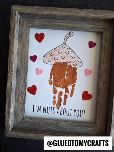 A framed Mother’s Day keepsake featuring a handprint painted as an acorn, surrounded by red and pink hearts, with the text “I’M NUTS ABOUT YOU!” at the bottom and @GLUEDTOMYCRAFTS in the corner.