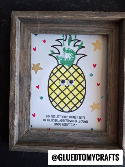 Framed art of a pineapple with a green handprint crown, googly eyes, and grid pattern. Decorated with heart, star, and gem stickers—this charming mother's day keepsake reads: “For the lady who is totally sweet… Happy Mother’s Day!”.