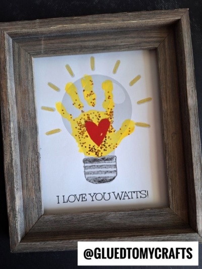 A framed artwork features a yellow handprint designed as a glowing lightbulb with a red heart in the center. Perfect as a Mother's Day keepsake, it reads "I LOVE YOU WATTS!" with the handle @GLUEDTOMYCRAFTS at the bottom.