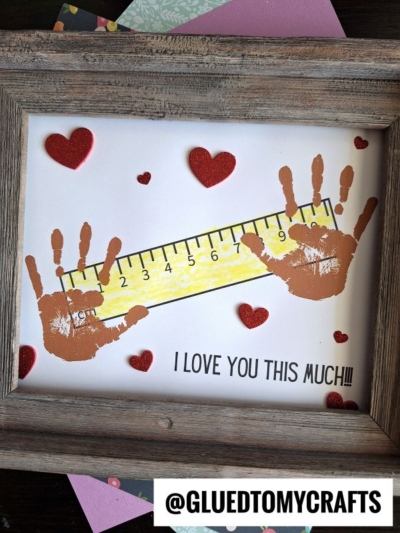 A framed mother’s day keepsake featuring two brown handprints on either side of a yellow ruler, surrounded by red hearts. Text below reads I LOVE YOU THIS MUCH!!! The handle @GLUEDTOMYCRAFTS is at the bottom.