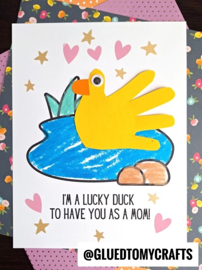 A handmade Mother’s Day keepsake card with a yellow duck made from a child’s handprint, blue pond, green reeds, and brown rocks. Pink hearts and gold stars decorate it with “I’M A LUCKY DUCK TO HAVE YOU AS A MOM!”.