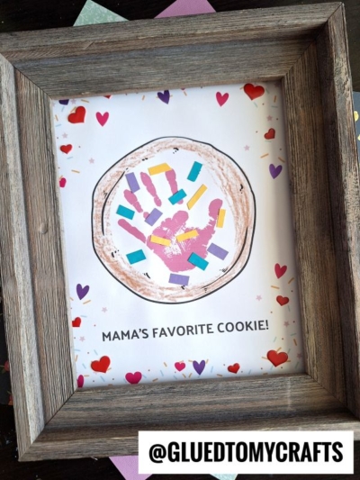 A framed mother’s day keepsake craft features a handprint decorated as a cookie with colorful paper pieces and hearts, surrounded by heart stickers. The text below reads “MAMA’S FAVORITE COOKIE!” with a @GLUEDTOMYCRAFTS label at the bottom.