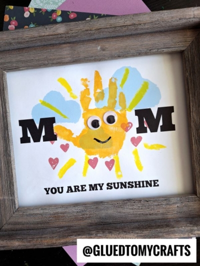 Framed craft with “MOM” text, a yellow handprint sun with googly eyes as the “O”, blue sky accents, pink hearts, and “YOU ARE MY SUNSHINE” below—a perfect mother's day keepsake. Instagram handle @GLUEDTOMYCRAFTS at the bottom.