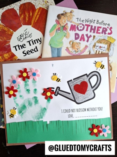 A handmade Mother's Day keepsake card with a green handprint as a flower stem, felt flowers, a watering can with a red heart, bee stickers, and a grass border. Two children's books and craft supplies are in the background.