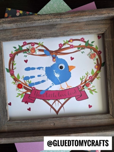 A unique mother's day keepsake: a framed craft with a blue handprint bird, googly eye, orange beak, and heart-shaped floral wreath. A pink banner reads "Your little love bird." @GLUEDTOMYCRAFTS is at the bottom.