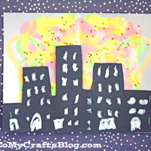 A craft project showing black paper cutouts of buildings with white windows against a colorful, paint splatter skyline background, all set on a star-patterned surface. The website GluedToMyCraftsBlog.com is at the bottom.