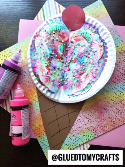 Colorful paint splat ice cream craft made from a painted paper plate, brown paper cone, and sprinkles, placed on bright patterned papers. Pink paint bottles are beside the craft. @GLUEDTOMYCRAFTS label is at the bottom.