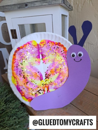 A colorful paint splat snails craft featuring a tie-dye coffee filter shell, gold glitter, and a smiling purple paper body with googly eyes. Instagram handle @GLUEDTOMYCRAFTS is shown at the bottom.
