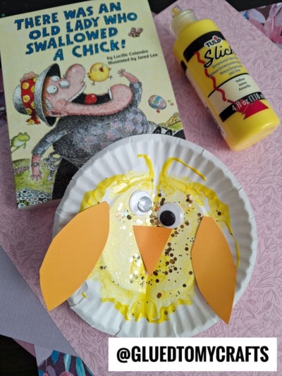 A children’s book titled “There Was an Old Lady Who Swallowed a Chick,” a bottle of yellow paint, and a paper plate baby chick craft with googly eyes and orange beak and wings sit on a pink floral background.