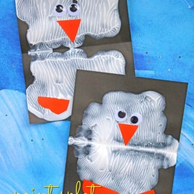 Two paint splat penguin crafts on dark paper, each with a white textured body, orange beak and feet, and googly eyes, displayed on a blue background. Text reads paint splat penguin or paint smash penguin at GluedToMyCraftsBlog.com.