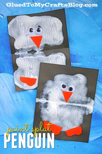 Two paint splat penguin crafts on dark paper, each with a white textured body, orange beak and feet, and googly eyes, displayed on a blue background. Text reads paint splat penguin or paint smash penguin at GluedToMyCraftsBlog.com.