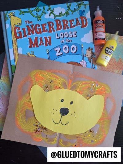A children’s book titled The Gingerbread Man Loose at the Zoo is shown with yellow and orange art supplies. In front is a paint splat lion face craft made of paper, paint, and glitter, with @GLUEDTOMYCRAFTS at the bottom.