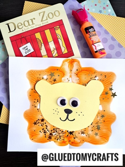 A children’s craft project of a paint splat lion’s face, made with orange paint, glitter, googly eyes, and yellow paper, sits next to a “Dear Zoo” book and orange glue on a colorful patterned background.