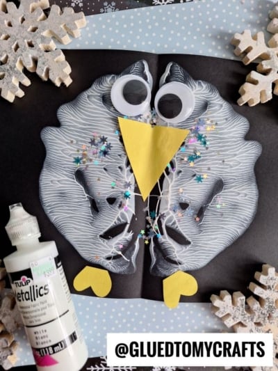 A winter owl craft made from a white paint symmetry print on black paper, similar to a paint splat penguin, decorated with googly eyes, a yellow beak, glitter, and surrounded by paper snowflakes and a bottle of white paint. Text reads @GLUEDTOMYCRAFTS.