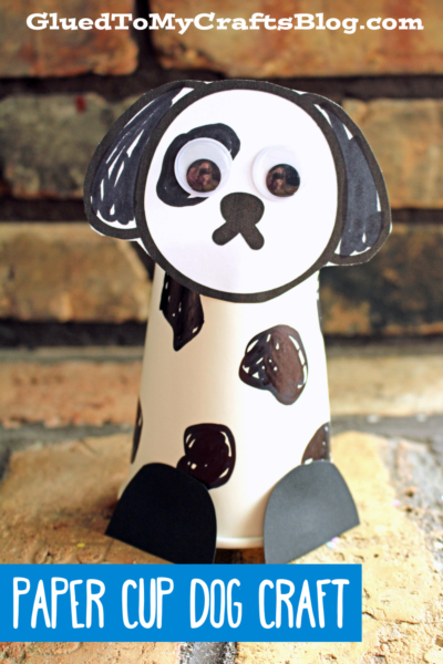 A paper cup dalmatian decorated with black paper ears, nose, spots, large googly eyes, and black paper paws. The background is a brick wall with text that reads Paper Cup Dog Craft.