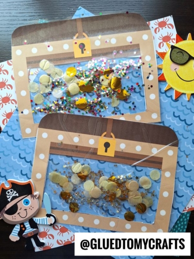 Two stained glass treasure chest crafts with clear windows hold sequins and confetti, placed on ocean-themed paper. A cartoon pirate and sun with sunglasses decorate the scene. @GLUEDTOMYCRAFTS is written at the bottom.