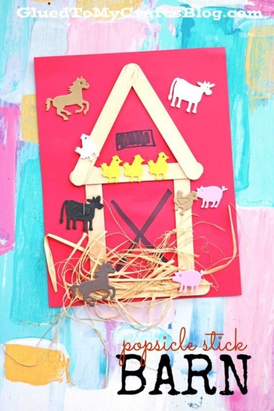 A charming popsicle stick barn stands on red paper, adorned with animal cutouts and real straw, all set against a vibrant, colorful painted background. Text reads: popsicle stick BARN.