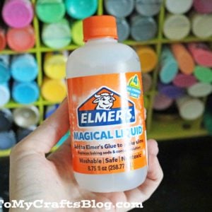 A hand holds a bottle of Elmers Magical Liquid slime activator in front of a shelf filled with colorful paint bottles. The label highlights it as safe, non-toxic, washable, and perfect for creating slime magical liquid fun.