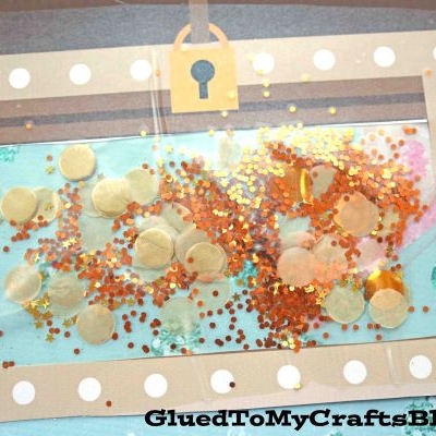 A stained glass treasure chest craft made with clear contact paper display sparkly confetti and gold sequins inside. The background features pastel starfish and sea-themed patterns.