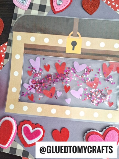 A handmade mailbox craft decorated with red, pink, and purple hearts and glitter, set on a background with layered felt hearts and a plaid pattern—like finding a stained glass treasure chest of love. The handle @GLUEDTOMYCRAFTS is at the bottom.