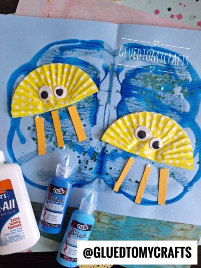 A cupcake liner jellyfish craft featuring two jellyfish made from yellow polka dot cupcake liners, googly eyes, and yellow paper strips on a blue painted background, with bottles of glue and glitter glue nearby.