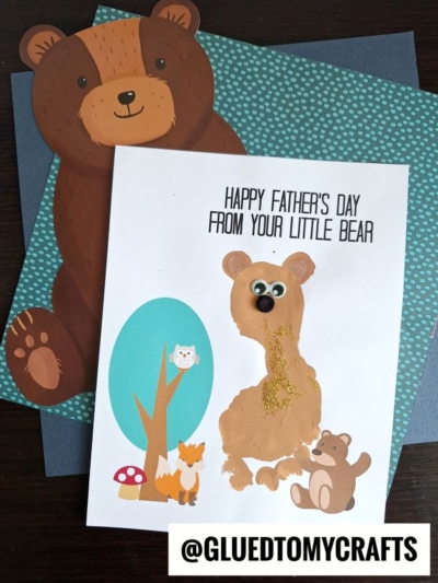 A handmade Father's Day card featuring a footprint baby bear made of paint handprints, a button nose, and gold glitter. Text reads "Happy Father's Day from your little bear." Background includes cartoon bears and a teal polka dot sheet.