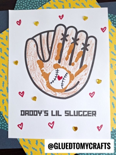 A craft featuring a handprint baseball mitt with a child’s handprint and a baseball in the center, “daddy’s lil slugger” written below, and small hearts all around. @GLUEDTOMYCRAFTS is at the bottom.