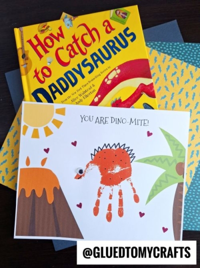 A children’s book titled “How to Catch a Daddysaurus” lies next to a handprint dino-mite keepsake card featuring an orange handprint dinosaur, palm trees, a sun, hearts, and the words “You are dino-mite!” @GLUEDTOMYCRAFTS.