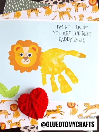 A children’s craft card features a handprint lion made from yellow paint, with a drawn face and mane. Text reads, IM NOT LION! YOU ARE THE BEST DADDY EVER! Decorative jungle-themed papers border this lion Father's Day keepsake.
