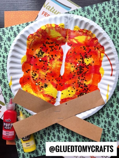 A paper plate decorated to look like a butterfly with red, yellow, and orange paint splat campfire colors, black glitter, and two brown paper strips for antennae. Bottles of paint and craft paper are nearby. @GLUEDTOMYCRAFTS is written at the bottom.
