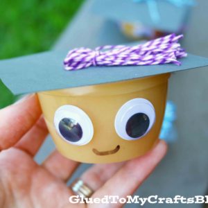 A hand holds a Graduation Applesauce Cup decorated as a graduation face, with googly eyes, a drawn smile, a black paper cap, and a purple-and-white tassel. The text GluedToMyCraftsBlog.com is at the bottom.