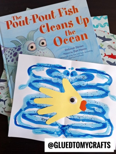 A children’s book titled “The Pout-Pout Fish Cleans Up the Ocean” sits beside a Paint Splat Ocean-themed fish craft, made from blue paint and a yellow handprint with a googly eye and orange lips. The image is labeled @GLUEDTOMYCRAFTS.
