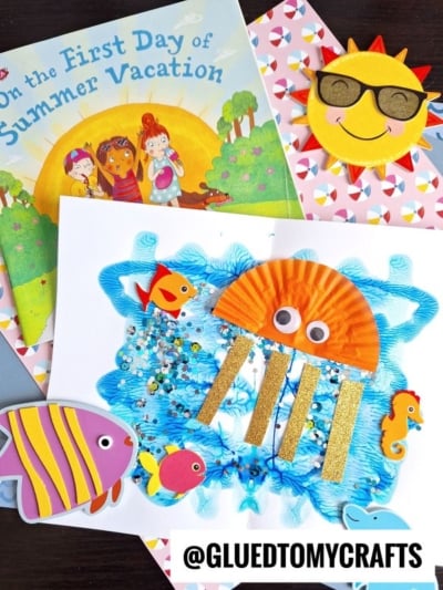 A colorful summer-themed craft scene with a children’s book, smiling sun cutout, cupcake liner jellyfish craft with glitter and googly eyes, plus fish and seahorse decorations. @GLUETOMYCRAFTS label at the bottom.