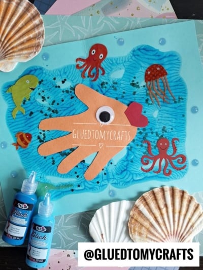 A colorful Paint Splat Ocean-themed craft features a handprint cutout as a fish with a red fin, surrounded by paper sea creatures, glitter, shells, and blue paint. GLUEDTOMYCRAFTS is written across the artwork.