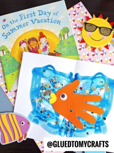 A children’s book titled On the First Day of Summer Vacation, a Paint Splat Ocean craft with a yellow sun in sunglasses, a fish craft, and a blue sensory bag with an orange handprint fish are arranged on a table. @GLUEDTOMYCRAFTS is written at the bottom.