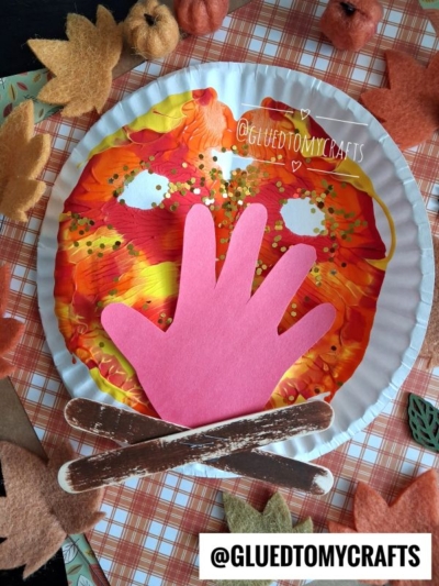 A paper plate painted with autumn colors using a paint splat technique forms a mask with eye holes, decorated with glitter. A pink paper hand and two brown craft sticks resemble a campfire, while felt leaves and plaid background enhance the fall theme.