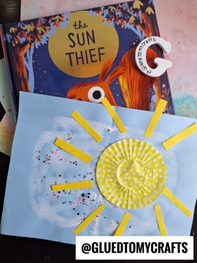 A sunshine craft for kids: a paint splat sun made from a yellow cupcake liner with yellow paper rays on blue paper, sitting atop The Sun Thief book. A clay crescent moon and glitters make it perfect for a sunny day craft project.