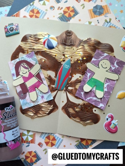 A handmade paint splat beach day card with two paper people, a surfboard, and beach stickers, surrounded by craft supplies and patterned papers. The username @GLUEDTOMYCRAFTS is displayed at the bottom.