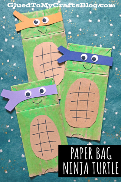 Three paper bag ninja turtles decorated with green paint, brown oval shells, and colorful paper masks sit on a blue background with white and gold star patterns. Text reads Paper Bag Ninja Turtle.