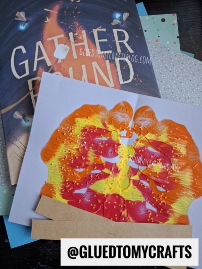 A colorful orange and yellow butterfly painting, created using the paint splat technique, sits atop a Gather Round book and polka dot paper. Text overlay reads @GLUEDTOMYCRAFTS.