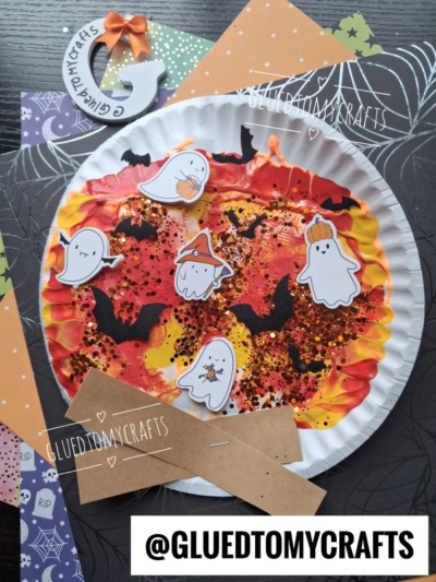 A Halloween-themed craft features a painted paper plate with ghosts, bats, and glitter on colorful Halloween papers, completed with a whimsical paint splat campfire effect for extra spookiness. A decorated G with a bow and an @GLUEDTOMYCRAFTS label are included.