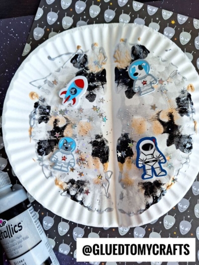 A paper plate moon decorated with black, white, and gold paint splat moon effects, space-themed stickers of rockets and astronauts, and metallic star confetti. Craft paint and a star-patterned background are visible. @GLUETOMYCRAFTS is written at the bottom.