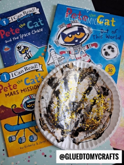 Three Pete the Cat space-themed books and a paper plate moon craft—painted black and yellow to resemble a paint splat moon—sit on a pink surface. A watermark reading @GLUEDTOMYCRAFTS appears twice on the image.