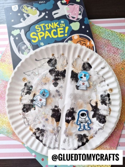 A paint splat moon paper plate decorated as a moon surface with cotton and astronaut stickers sits on colorful paper next to the “Stink in Space!” children’s book. @GLUEDTOMYCRAFTS is written at the bottom.