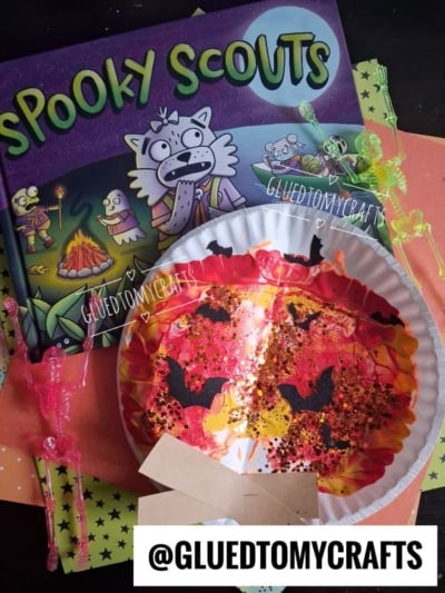 A children’s book titled Spooky Scouts, a painted paper plate craft with black and orange glitter, colorful skeleton straws, and orange polka-dot paper, featuring a whimsical paint splat campfire design. @GLUEDTOMYCRAFTS watermark is visible.